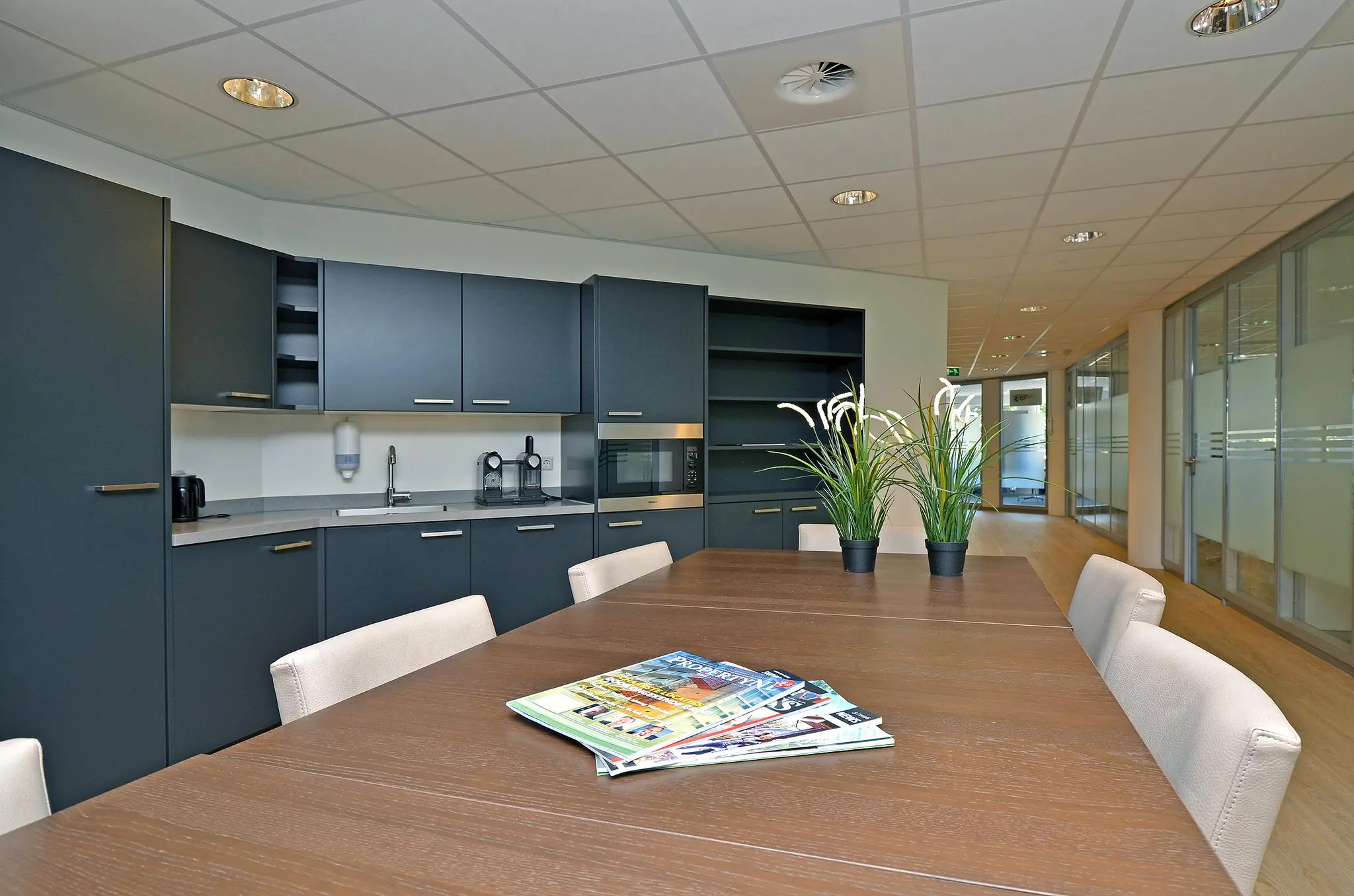 Moderne kantoorkeuken met een vergadertafel, grijze kasten, planten en tijdschriften op tafel.